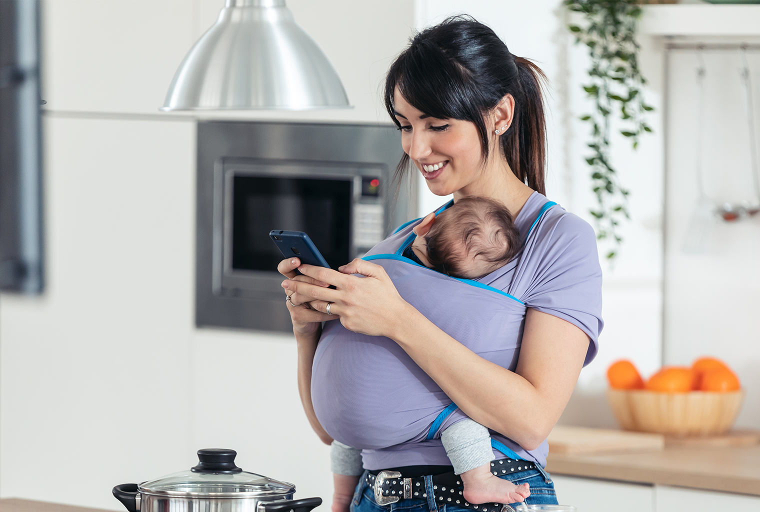 young mom holding baby in sling and cell phone in her hands