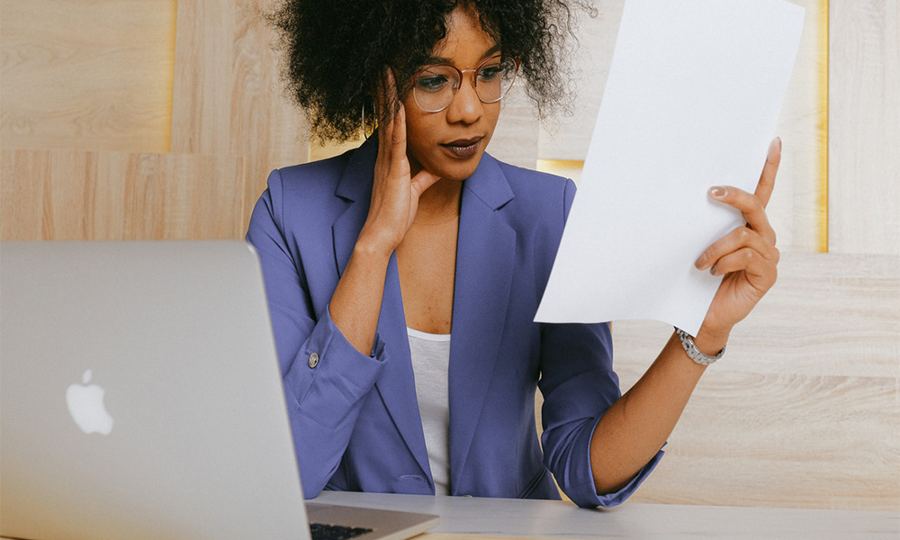 Young Woman Wearing Glasses Sitting At Desk Looking At Paper